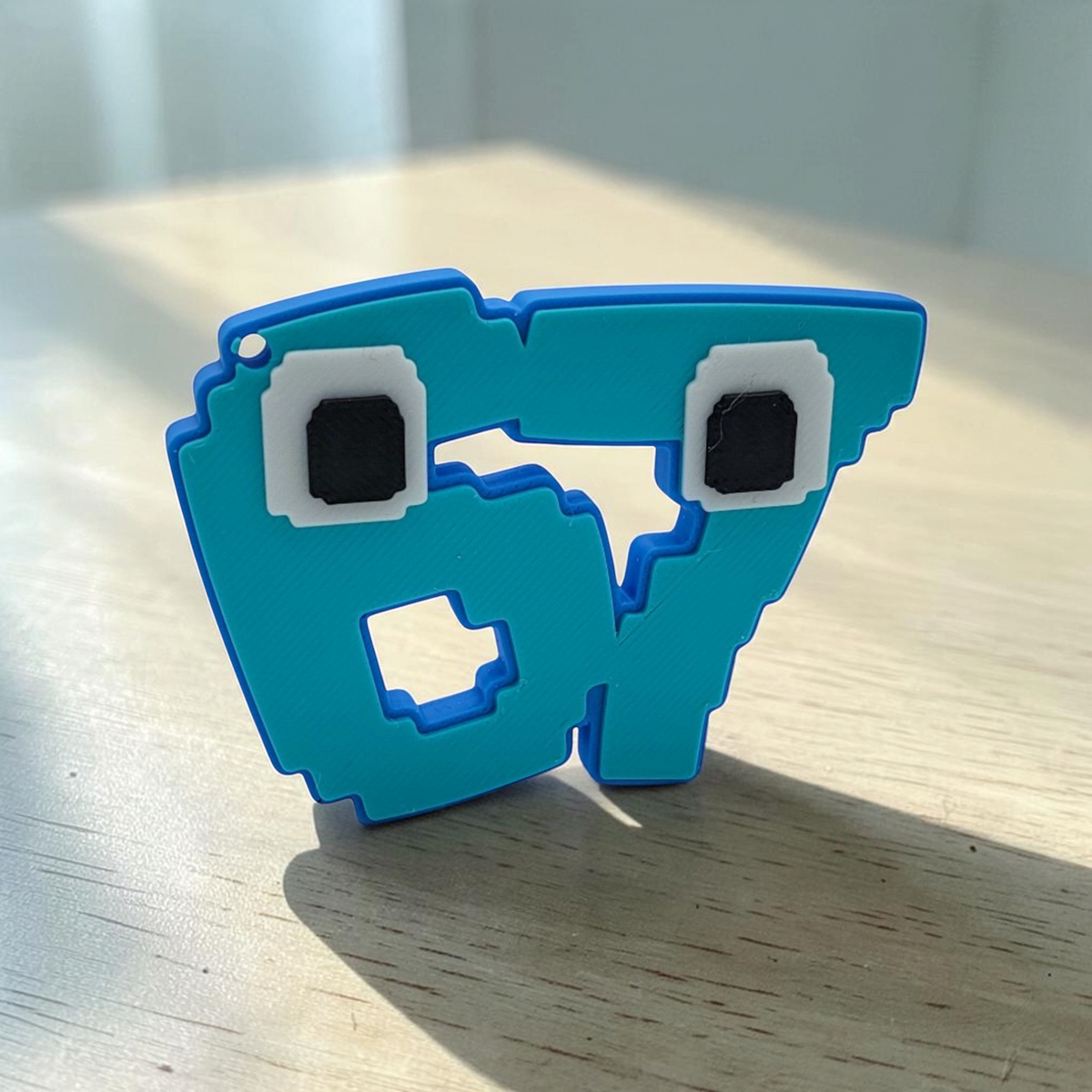 Front view of a blue pixelated 67 meme character keyring with square eyes on a white background.