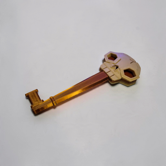 Gold and bronze colored 3D printed skeleton key with a skull shaped bow displayed on a textured stone surface.