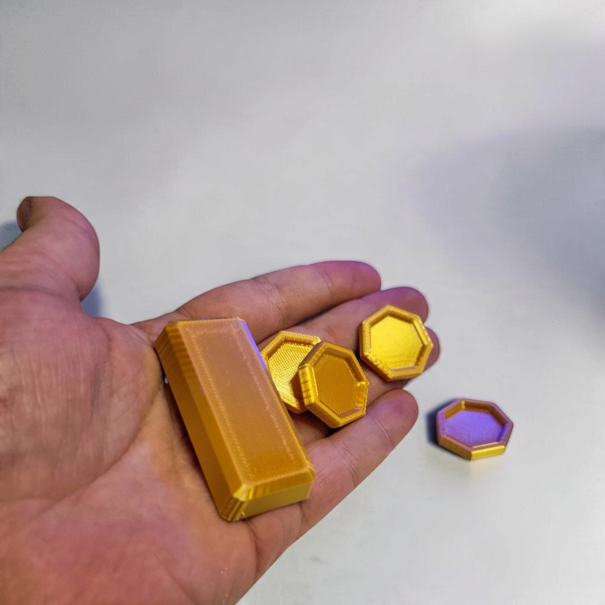 A hand holds the gold colored hexagonal coins and rectangular bar to show the size of the 3D printed set.
