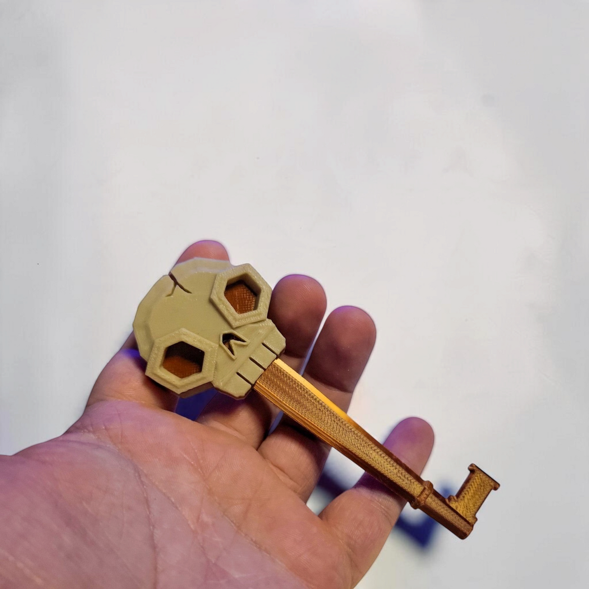 A hand holds the gold and bronze skeleton key, showing its long decorative shaft and skull shaped bow.