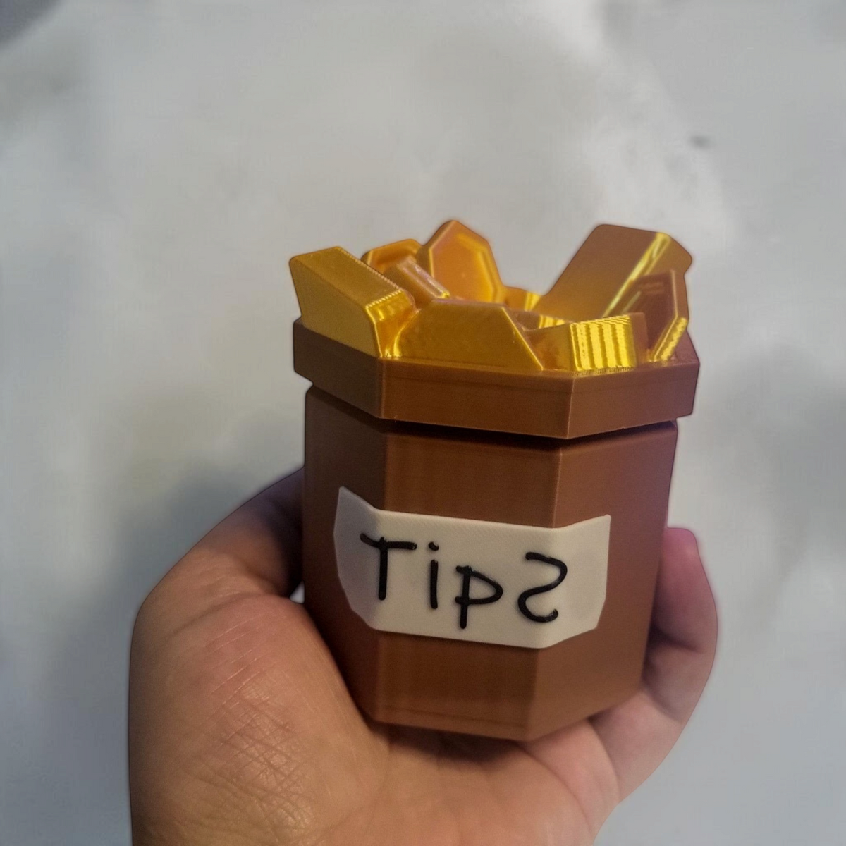 A hand holds the bone and copper 3D printed tip jar, showing its handwritten Tips label and faceted design.