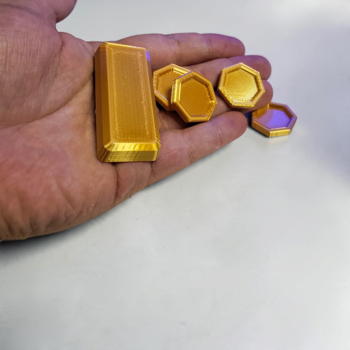 Gold colored 3D printed hexagonal coins and a rectangular bar displayed in an open palm against a light background.
