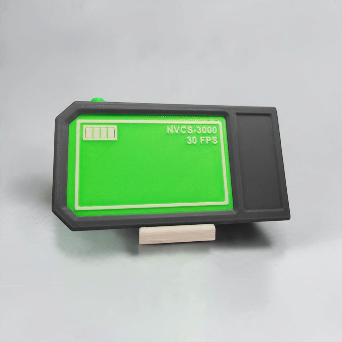 Front view of the gray NVCS 3000 handheld prop with green display, red and green buttons, and small white stand.