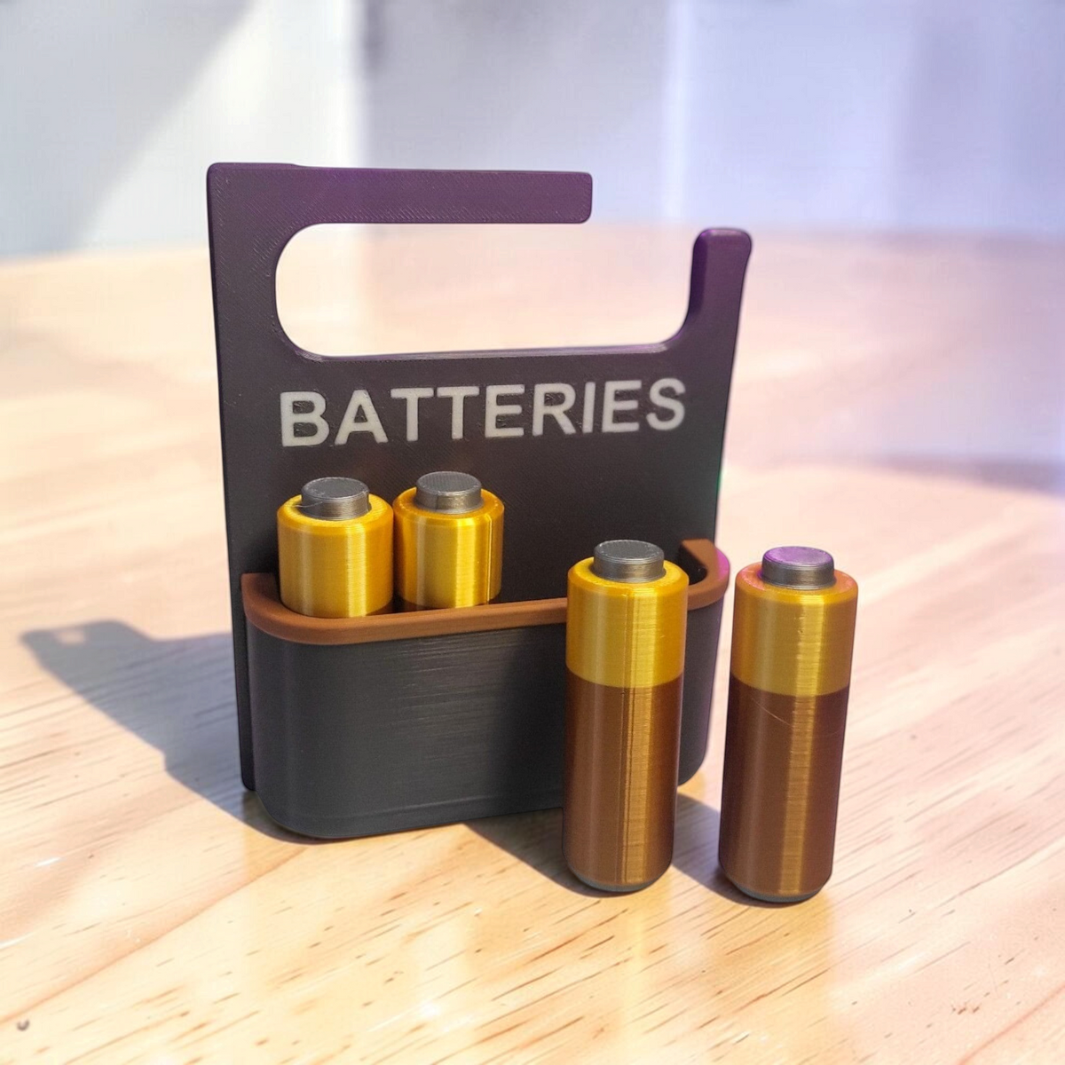 Black 3D-printed battery holder labelled “BATTERIES”, with two gold-coloured AA batteries displayed in front.