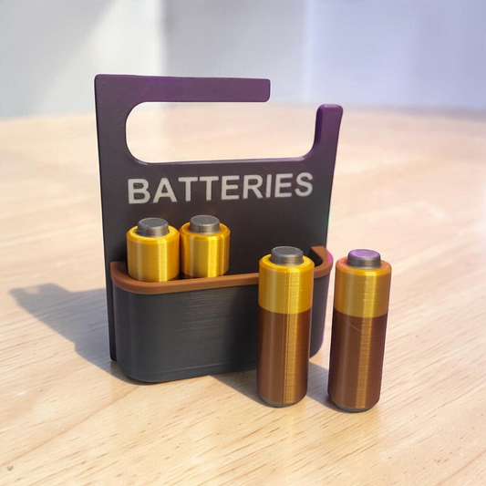 Black 3D-printed battery holder labelled “BATTERIES”, displaying gold and copper-coloured AA batteries on a desk.