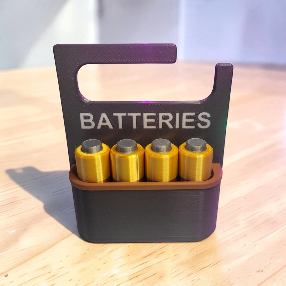 Front view of a black 3D-printed battery organiser labelled “BATTERIES”, holding four gold-coloured AA batteries.